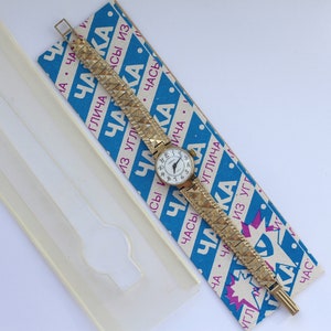 May include: A gold-tone wristwatch with a white face and black Roman numeral hour markers. The watch has a gold-tone metal band with a textured pattern. The watch is on a blue and white striped box with the word "ЧАККА" printed on it.