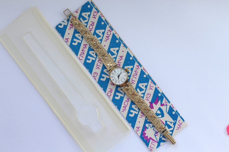 May include: A gold-tone wristwatch with a white face and black Roman numeral hour markers. The watch has a gold-tone metal band with a textured pattern. The watch is on a blue and white striped box with the word "ЧАККА" printed on it.