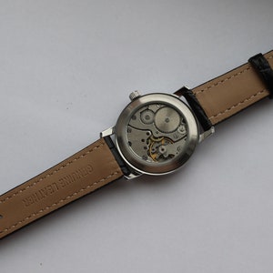 May include: A silver wristwatch with a brown leather band. The back of the watch is open, revealing the inner workings of the watch.