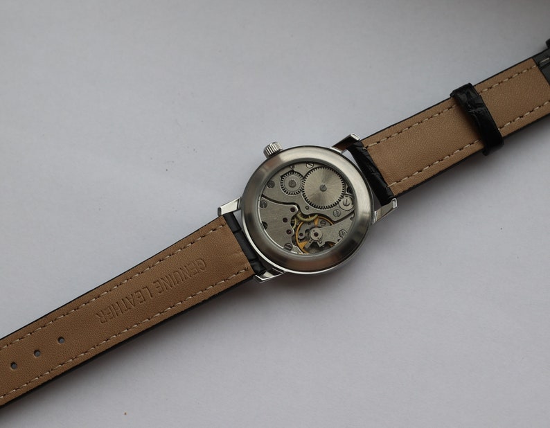 May include: A silver wristwatch with a brown leather band. The back of the watch is open, revealing the inner workings of the watch.