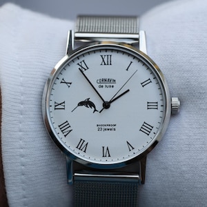 May include: A silver watch with a white face and black Roman numeral hour markers. The watch has a silver mesh band and a small dolphin logo on the face. The text "CORNAVIN de Luxe" and "Shockproof 23 Jewels" are printed on the face.