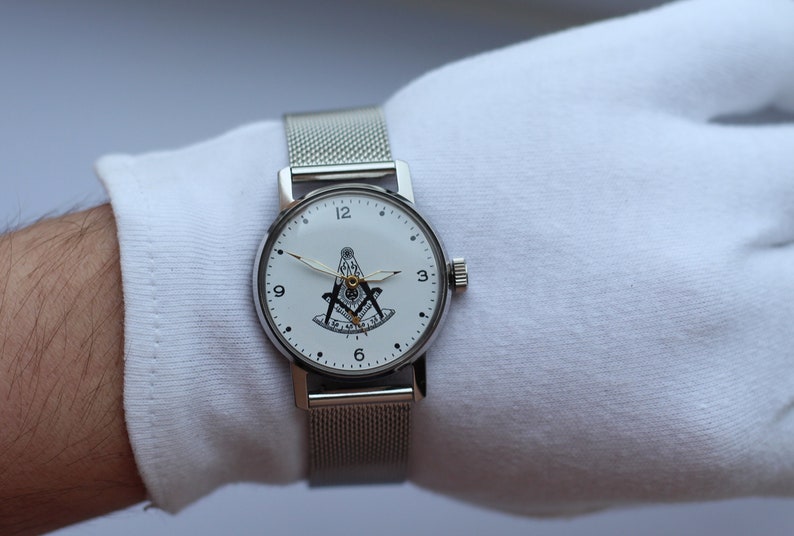 May include: A silver watch with a white face and a silver mesh band. The face of the watch features a black and white image of a compass and square, a symbol of Freemasonry.