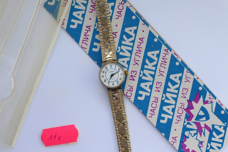May include: A gold-tone wristwatch with a white face and black Roman numeral hour markers. The watch has a gold-tone metal band with a textured pattern. The watch is on a blue and white box with the text "ЧАЙКА" in pink and blue.