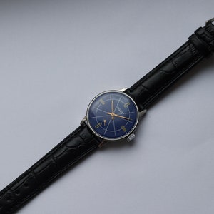 Puede incluir: Un reloj de pulsera vintage con una esfera azul y detalles dorados. El reloj tiene una correa de cuero negro con cierre de hebilla.