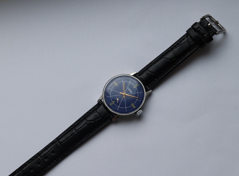 Puede incluir: Un reloj de pulsera vintage con una esfera azul y detalles dorados. El reloj tiene una correa de cuero negro con cierre de hebilla.