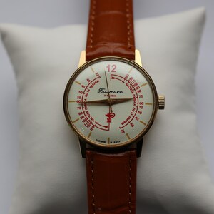 May include: A vintage gold-toned wristwatch with a white face and red markings, including the number 12. The watch has a brown leather band and is displayed on a white pillow. The watch face has Cyrillic text.