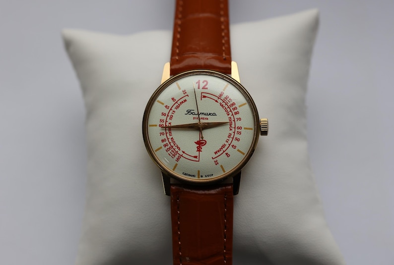 May include: A vintage gold-toned wristwatch with a white face and red markings, including the number 12. The watch has a brown leather band and is displayed on a white pillow. The watch face has Cyrillic text.