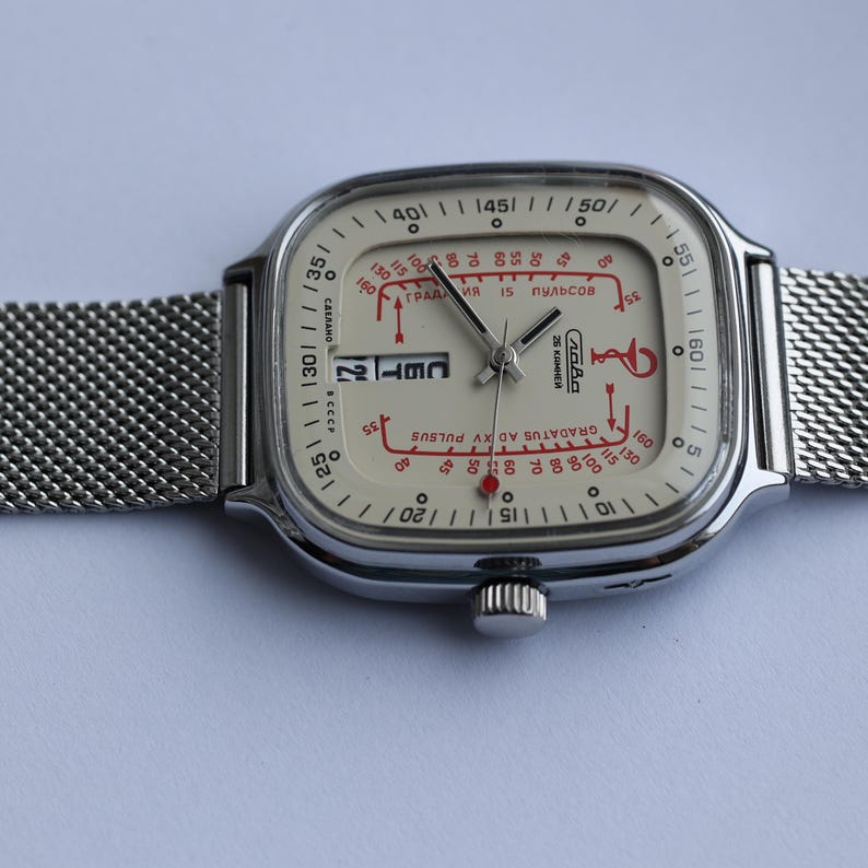 May include: A vintage silver-toned wristwatch with a square face and a mesh band. The watch face is off-white with red and black markings, including the date. The watch has a retro design.
