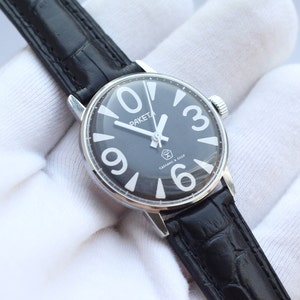 Puede incluir: Un reloj de pulsera con esfera negra, números arábigos blancos y una correa de cuero negra. La esfera del reloj dice "ПАКЕТА" y "СССР".
