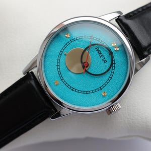 May include: A silver wristwatch with a black leather band and a turquoise blue face. The face has a gold circle with a red hand and the word "PAKETA" printed on it.