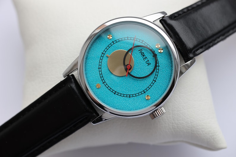 May include: A silver wristwatch with a black leather band and a turquoise blue face. The face has a gold circle with a red hand and the word "PAKETA" printed on it.