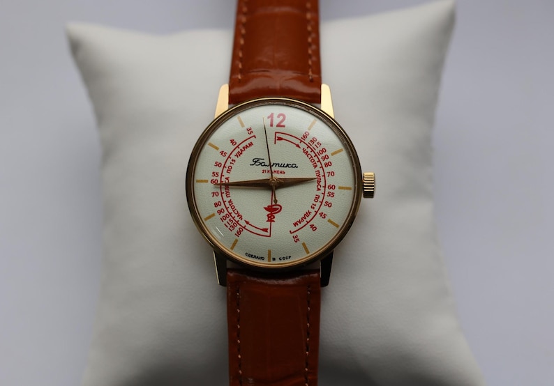 May include: A vintage gold-toned wristwatch with a cream-colored face and brown leather strap. The watch face features red markings and the word "Baltika". The watch is displayed on a white pillow.