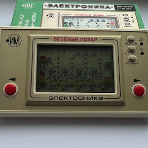 Puede incluir: Un juego electrónico portátil vintage con una carcasa beige, botones rojos y una pequeña pantalla que muestra un juego de cocina. El juego está etiquetado como "Vesely Povar" y "Elektronika". Un folleto verde es visible.