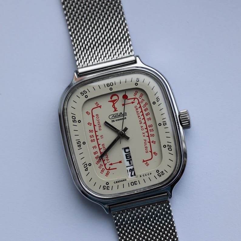 May include: A vintage silver-toned wristwatch with a square face and a mesh band. The watch face is off-white with black hands and red accents, featuring text and a date display. The watch has a retro aesthetic.