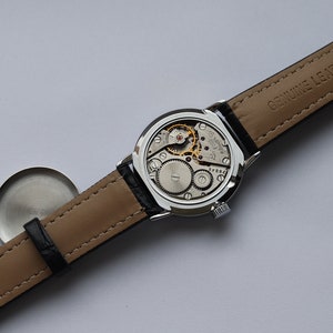 Puede incluir: Un reloj de pulsera plateado con una correa de cuero negro. La esfera del reloj est&aacute; expuesta, mostrando el mecanismo interno del reloj. La correa est&aacute; etiquetada como "Genuine Leather".