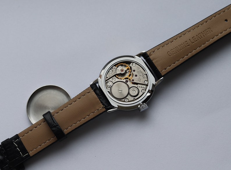 Puede incluir: Un reloj de pulsera plateado con una correa de cuero negro. La esfera del reloj est&aacute; expuesta, mostrando el mecanismo interno del reloj. La correa est&aacute; etiquetada como "Genuine Leather".