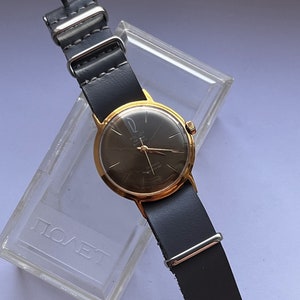 May include: A gold-tone wristwatch with a black face and a black leather strap. The watch face has the words "POLJOT" and "21 jewels" printed on it. The watch is in a clear plastic case with the word "ПОЛЕТ" printed on it.
