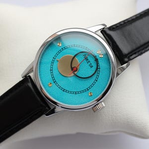 May include: A silver wristwatch with a black leather band. The watch face is turquoise blue with gold accents and the word "PAKE TA" printed on it.