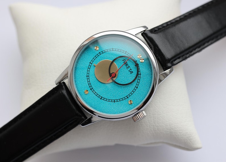 May include: A silver wristwatch with a black leather band. The watch face is turquoise blue with gold accents and the word "PAKE TA" printed on it.