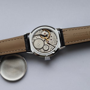 Puede incluir: Un reloj de pulsera plateado con una correa de cuero negro. La esfera del reloj es visible y muestra el funcionamiento interno del reloj. La correa est&aacute; etiquetada como "GENUINE LEATHER".