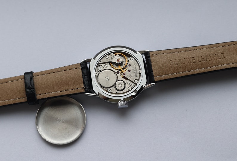 Puede incluir: Un reloj de pulsera plateado con una correa de cuero negro. La esfera del reloj es visible y muestra el funcionamiento interno del reloj. La correa est&aacute; etiquetada como "GENUINE LEATHER".