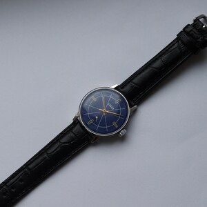 Puede incluir: Un reloj de pulsera vintage con una esfera azul y detalles dorados. El reloj tiene una correa de cuero negro con un patr&oacute;n de cocodrilo.