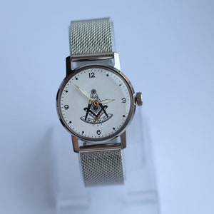 May include: A silver watch with a white face and a silver mesh band. The face of the watch has black Roman numeral hour markers and a black and gold Masonic symbol in the center.