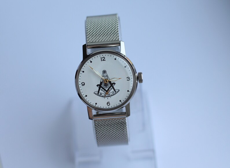 May include: A silver watch with a white face and a silver mesh band. The face of the watch has black Roman numeral hour markers and a black and gold Masonic symbol in the center.