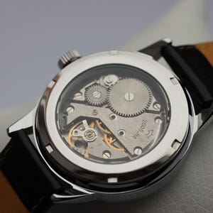 May include: A silver wristwatch with a black leather band and a clear case back revealing the inner workings of the watch. The watch face has the text "ЧЧ 2609" printed on it.