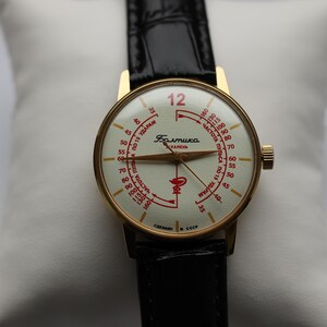 May include: Vintage gold-tone wristwatch with a cream-colored face and black leather band. The watch face features red markings and text, including the number 12. The watch is displayed on a white pillow.