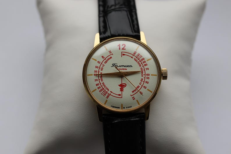 May include: Vintage gold-tone wristwatch with a cream-colored face and black leather band. The watch face features red markings and text, including the number 12. The watch is displayed on a white pillow.