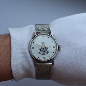 May include: A silver wristwatch with a white face and a black and gold Masonic symbol. The watch has a silver mesh band.