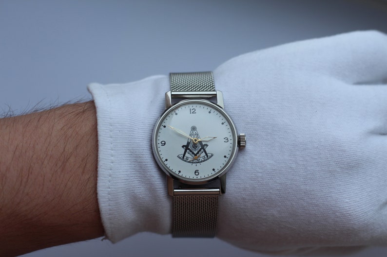 May include: A silver wristwatch with a white face and a black and gold Masonic symbol. The watch has a silver mesh band.