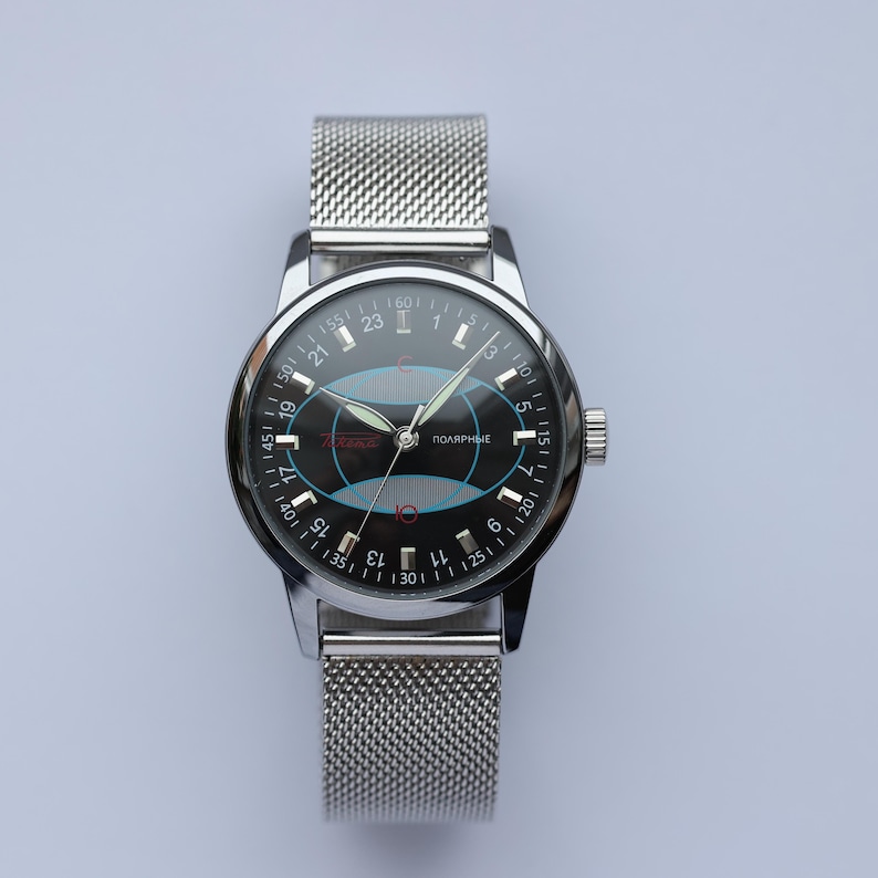 May include: A silver wristwatch with a mesh band and a black face. The watch face features a blue and red design with the word "ПОЛЯРНЫЕ" and white numerals. The watch has a silver bezel and a crown on the right side.