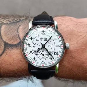 May include: A white wristwatch with black hands and a black leather band. The watch face has black Chinese characters and a black image of a building with birds flying around it.