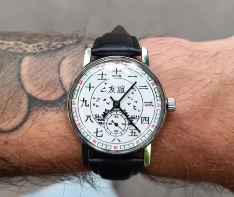 May include: A white wristwatch with black hands and a black leather band. The watch face has black Chinese characters and a black image of a building with birds flying around it.