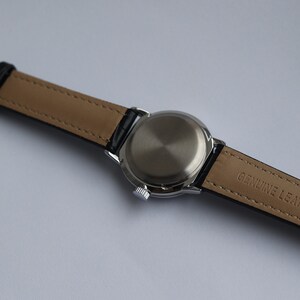 Puede incluir: Un reloj plateado con una correa de cuero negro. La correa est&aacute; etiquetada como "GENUINE LEATHER".