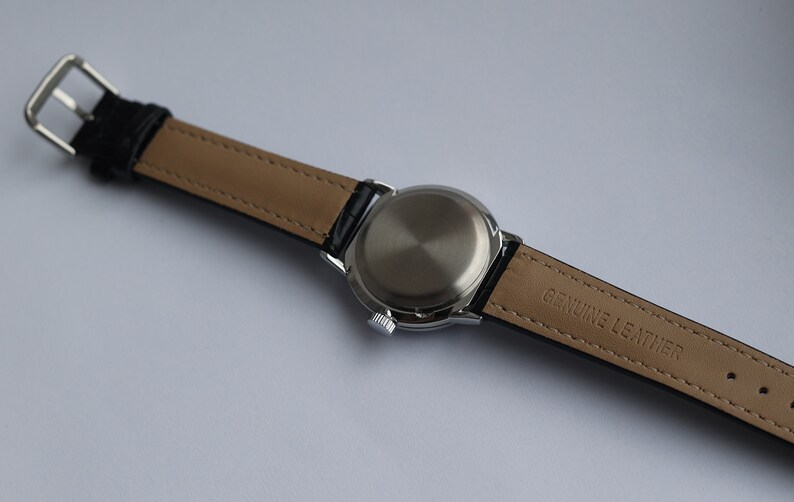 Puede incluir: Un reloj plateado con una correa de cuero negro. La correa est&aacute; etiquetada como "GENUINE LEATHER".
