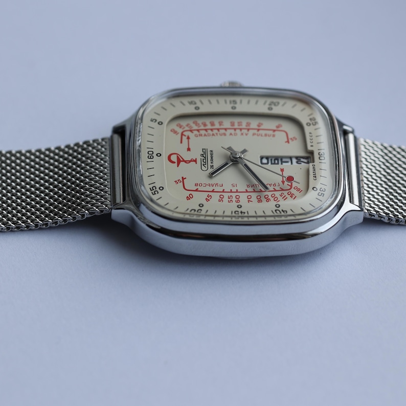 May include: A vintage silver-toned wristwatch with a square face and a mesh band. The watch face is cream-colored with red markings and text, including "GRADATUS AD KY PULSUS" and "15 PULSOS". The watch displays the date.