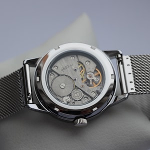 May include: Close-up of a silver wristwatch with a transparent back, revealing the intricate mechanical movement. The watch features a mesh band and a round face. The gears and springs are visible, showcasing the watch's internal components.