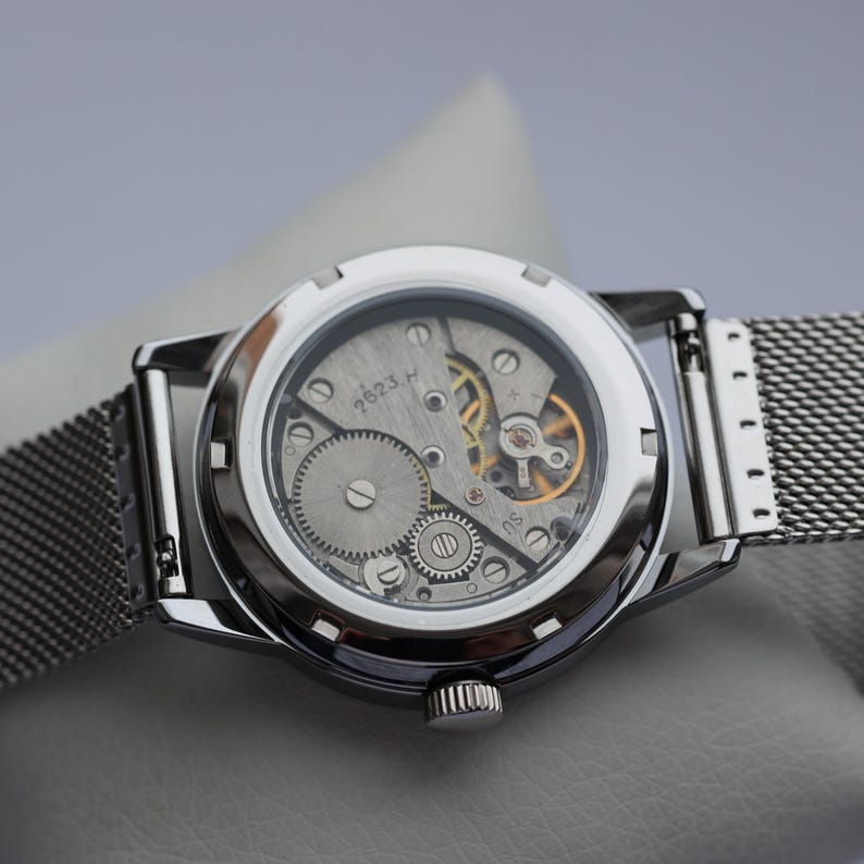 May include: Close-up of a silver wristwatch with a transparent back, revealing the intricate mechanical movement. The watch features a mesh band and a round face. The gears and springs are visible, showcasing the watch's internal components.