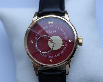 May include: A gold-tone wristwatch with a red face and a black leather band. The watch face features a white circle with the word "PAKE TA" printed inside.