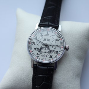 May include: A silver wristwatch with a white face and black leather band. The face has Chinese characters and a small image of a building with birds flying around it.