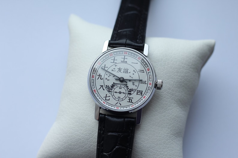 May include: A silver wristwatch with a white face and black leather band. The face has Chinese characters and a small image of a building with birds flying around it.