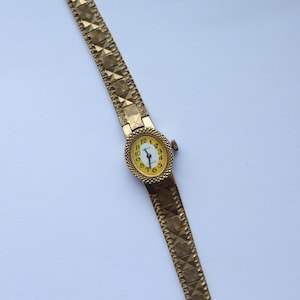May include: A gold-tone wristwatch with a yellow face and black Roman numeral hour markers. The watch has a gold-tone metal band with a textured pattern.