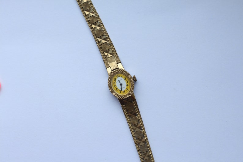May include: A gold-tone wristwatch with a yellow face and black Roman numeral hour markers. The watch has a gold-tone metal band with a textured pattern.