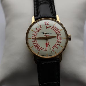 May include: A vintage gold-tone wristwatch with a cream-colored face and black leather band. The watch face features red numerals and text, including the number 12 in red. The watch is displayed on a white pillow.