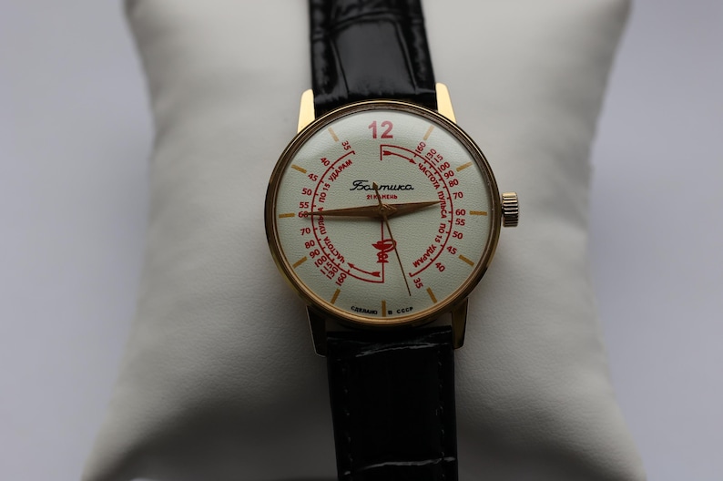 May include: A vintage gold-tone wristwatch with a cream-colored face and black leather band. The watch face features red numerals and text, including the number 12 in red. The watch is displayed on a white pillow.