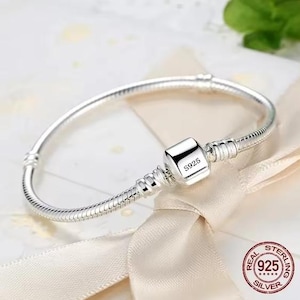 May include: A silver snake chain bracelet featuring a smooth, square-shaped clasp with the engraving S925. The bracelet has a flexible, rounded link design with two stationary threaded stations on either side of the central clasp mechanism.