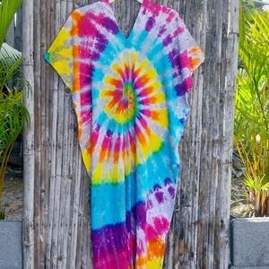 May include: A vibrant tie-dye caftan in a spiral pattern of yellow, orange, pink, purple, blue, and white. The V-neck garment is hanging on a white hanger against a bamboo fence. The colorful, flowing dress is a bohemian style.